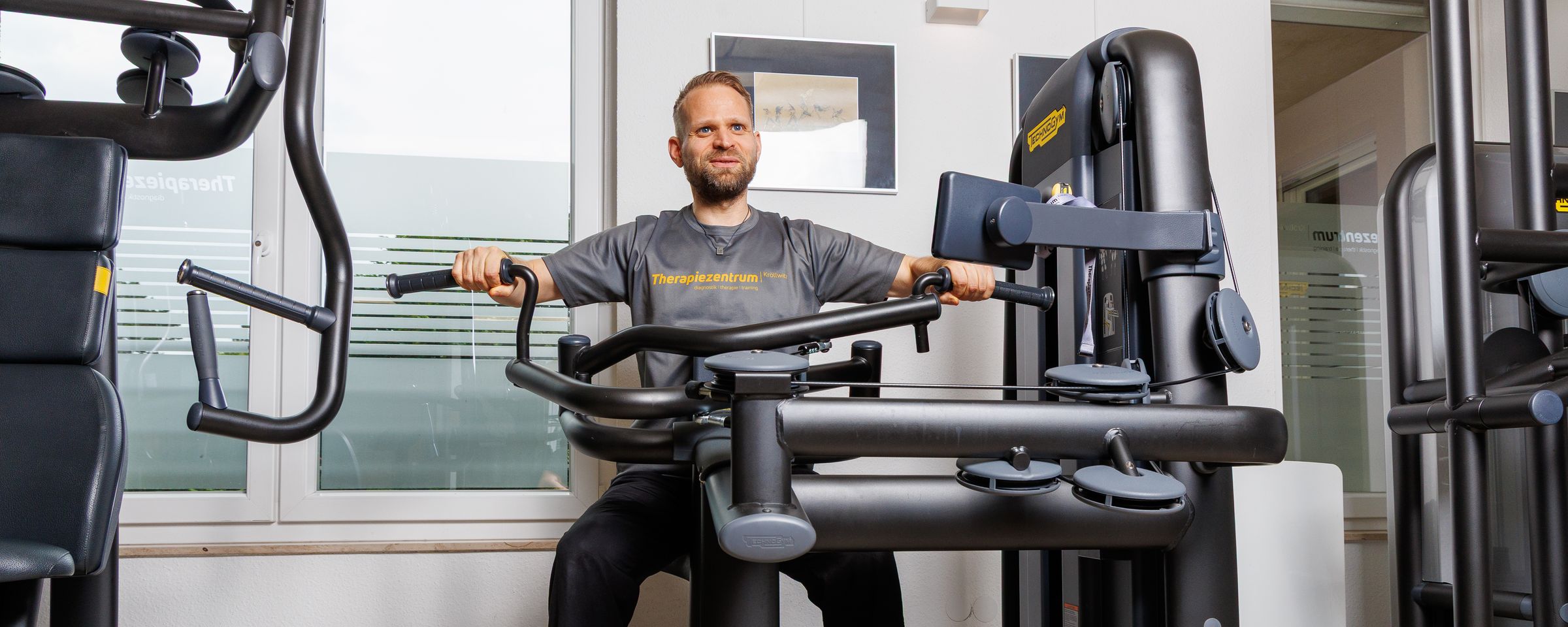 Trainer im Praxisshirt an einem Technogym-Gerät auf der Trainingsfläche