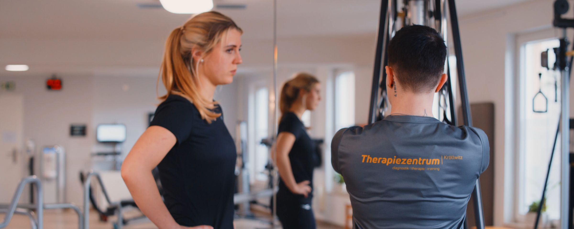 Patientin und Therapeut im Praxisshirt auf der Trainingsfläche, im Hintergrund eine weitere Patientin